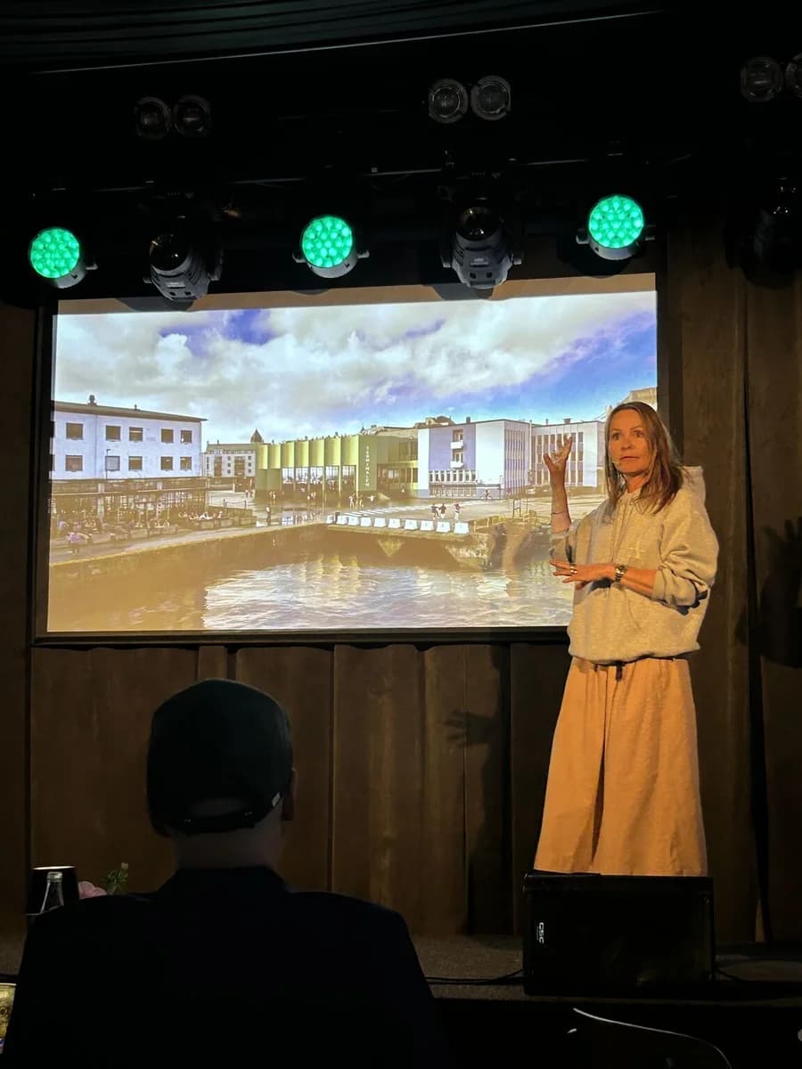 En foredragsholder på scenen foran et bilde av bygninger.